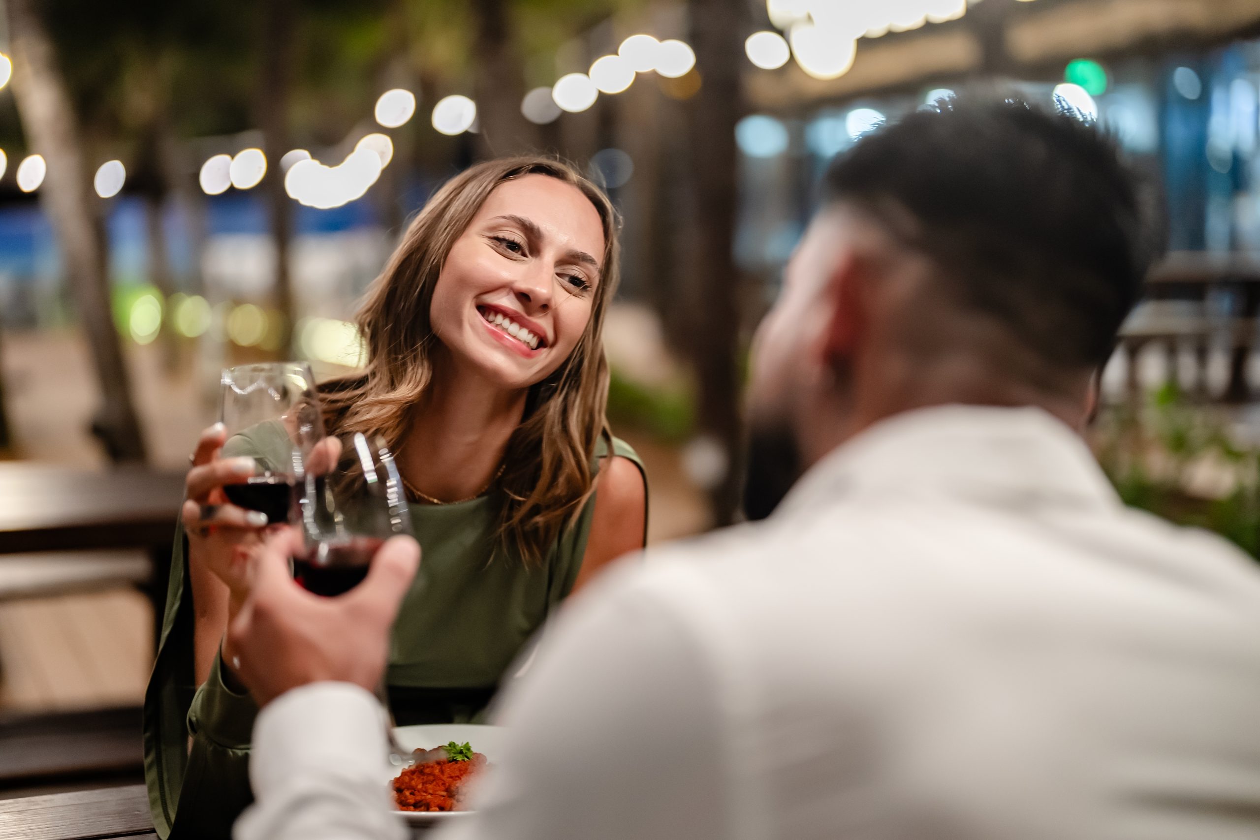 A couple enjoying a dinner date with glasses of red wine, smiling at each other in a cozy outdoor setting.