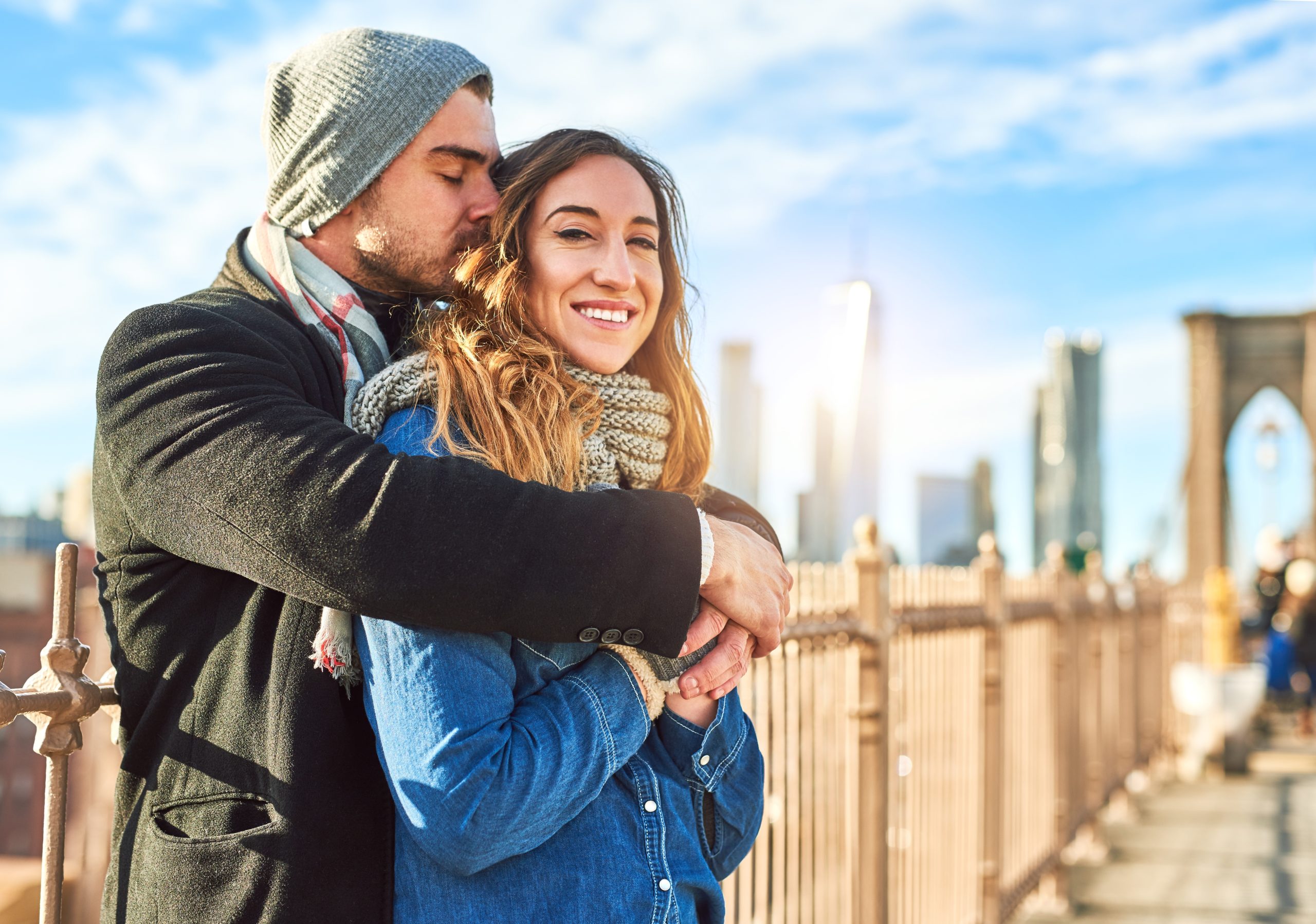 Couple embracing on a bridge with a city skyline in the background, smiling and enjoying a sunny day together.