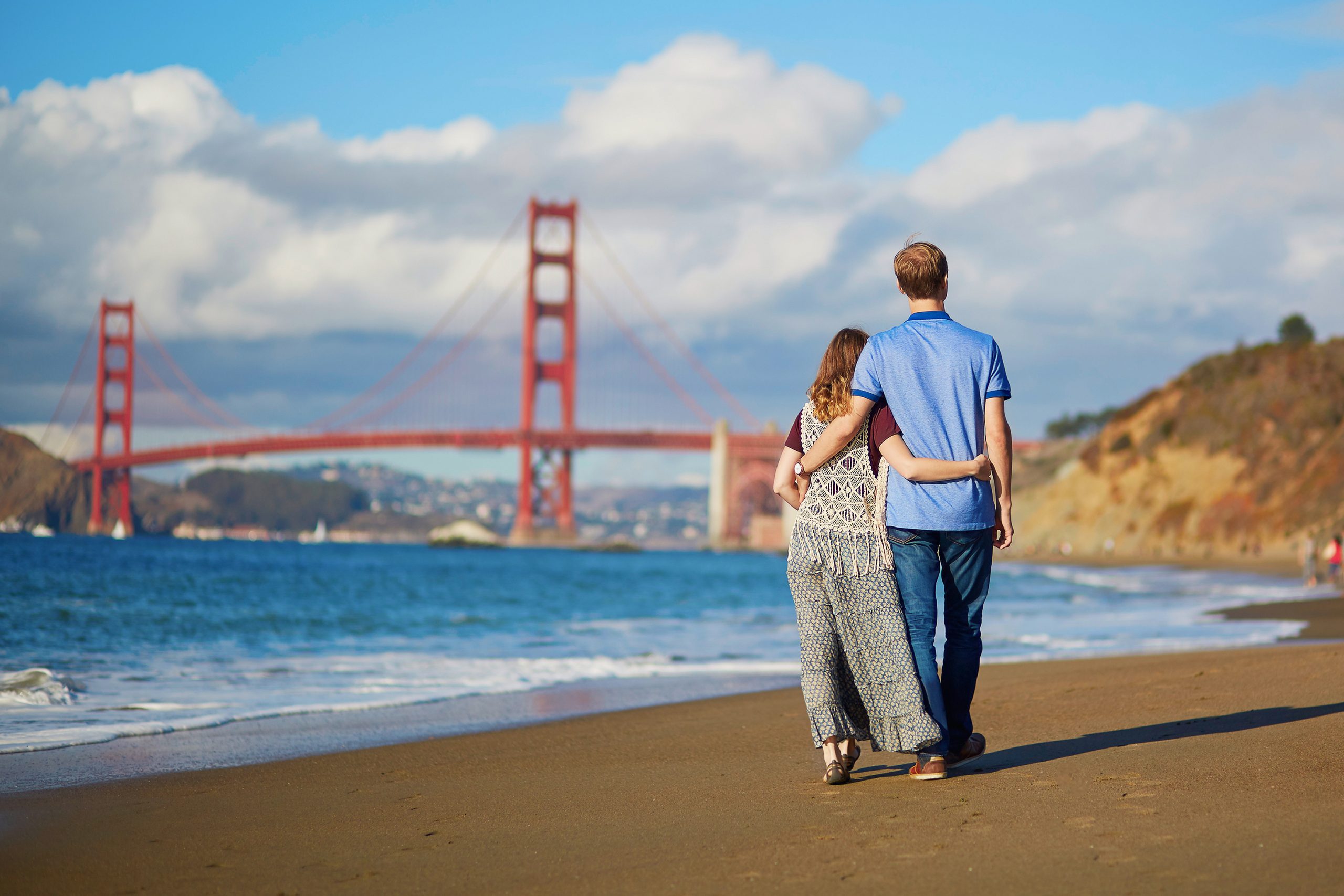 Couple walking along the beach with the Golden Gate Bridge in the background on a sunny day.
