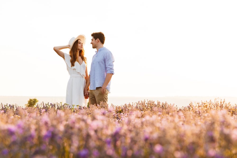 A couple stands in a lavender field, enjoying each other's company under a bright sky.