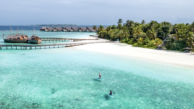 Two paddleboarders enjoy clear turquoise waters near tropical palm trees and overwater bungalows.