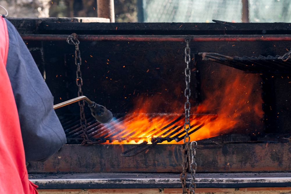 A person using a torch to light a barbecue grill with flames and smoke rising from the grates.