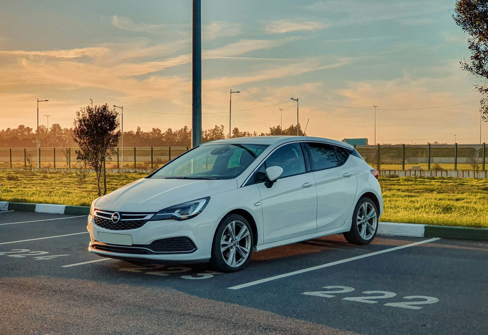 A white sedan parked in a parking space with a sunset backdrop and greenery in the background.