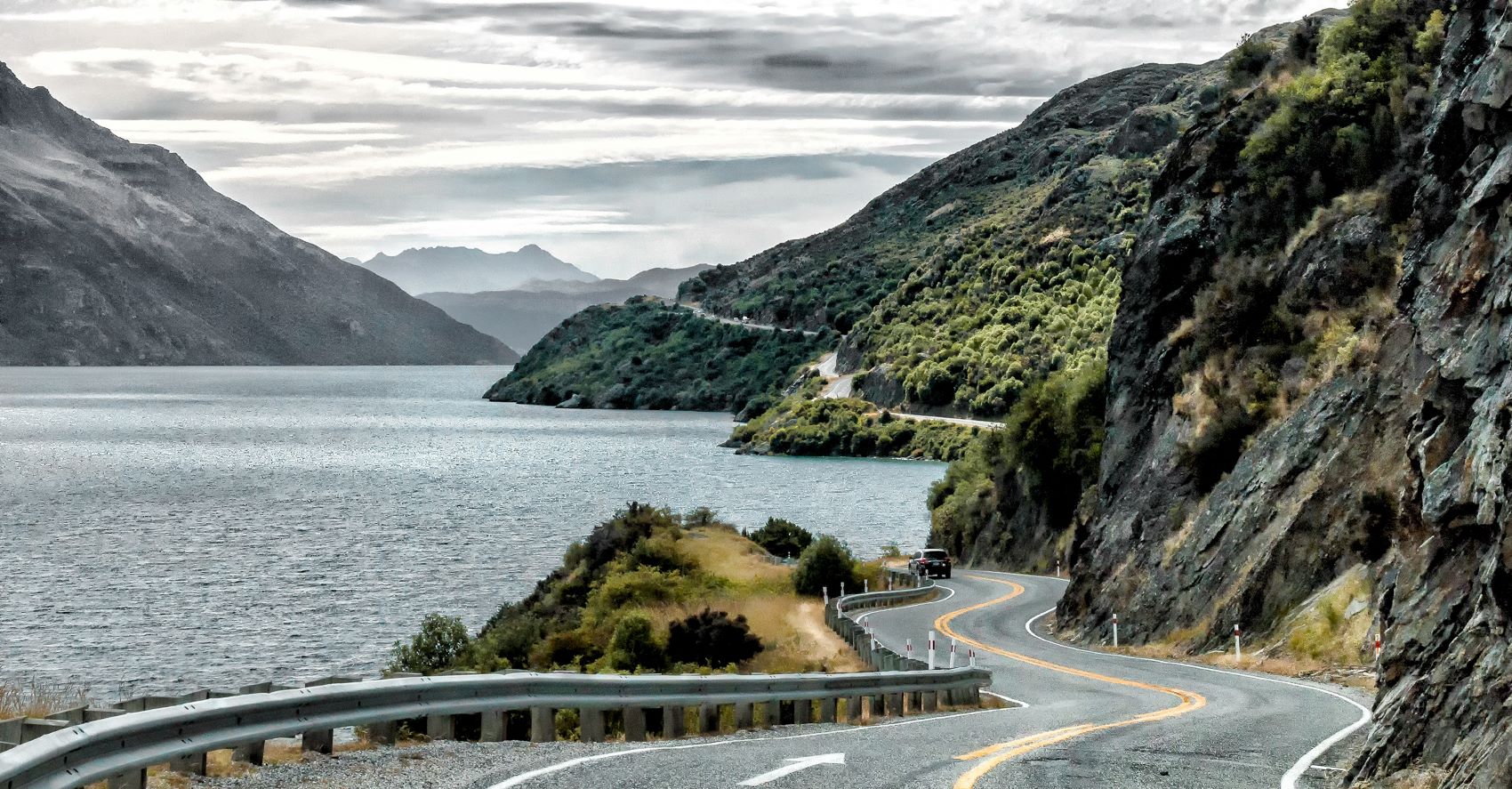 Winding road along a lake surrounded by mountains in New Zealand, highlighting a premium car rental experience.