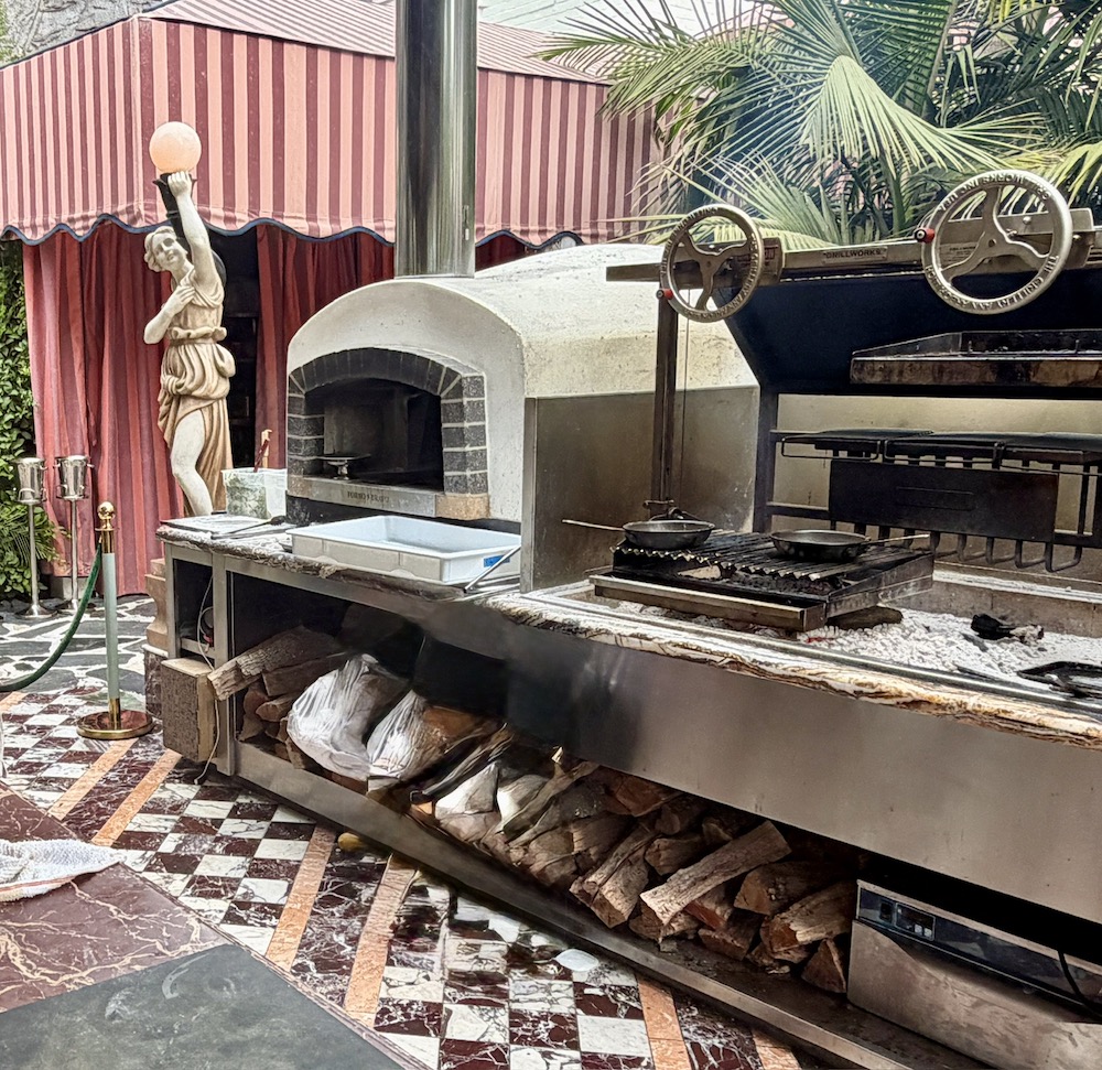 A grand outdoor oven setup with a stone pizza oven and grilling space, surrounded by a tropical-themed patio and statue.
