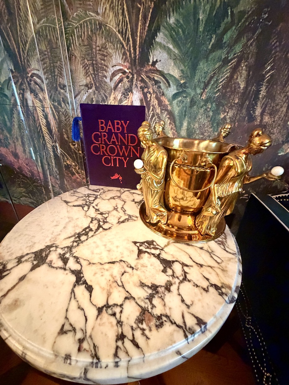Gold trophy-style vase next to a menu for Baby Grand Crown City on a marble table, with tropical wallpaper in the background.