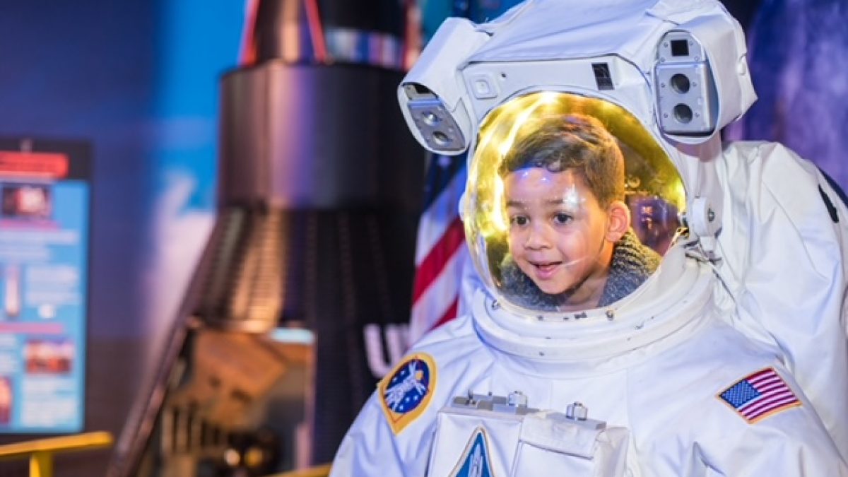A child smiles while wearing an astronaut suit at WonderWorks Myrtle Beach, with a rocket display in the background.