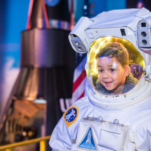 A child smiles while wearing an astronaut suit at WonderWorks Myrtle Beach, with a rocket display in the background.