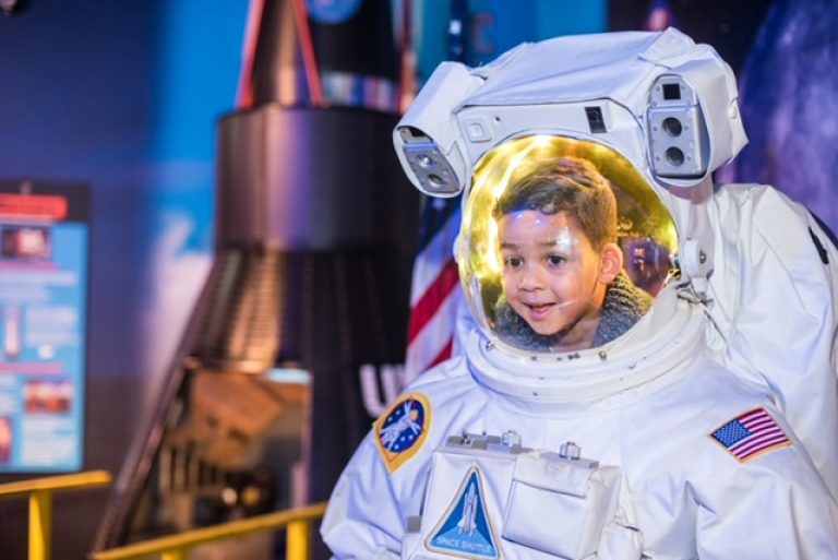 A child smiles while wearing an astronaut suit at WonderWorks Myrtle Beach, with a rocket display in the background.