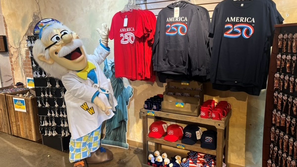 A character in a lab coat stands beside T-shirts and hats celebrating America’s 250th anniversary in a vibrant shop display.