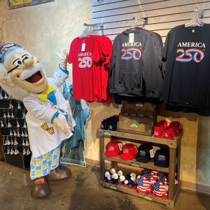 A character in a lab coat stands beside T-shirts and hats celebrating America’s 250th anniversary in a vibrant shop display.