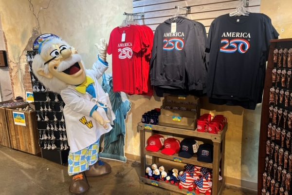 A character in a lab coat stands beside T-shirts and hats celebrating America’s 250th anniversary in a vibrant shop display.