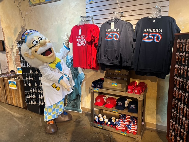 A character in a lab coat stands beside T-shirts and hats celebrating America’s 250th anniversary in a vibrant shop display.