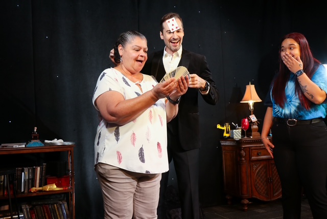 A magician performs with a card for an audience member, while another audience member reacts in surprise during a magic show.