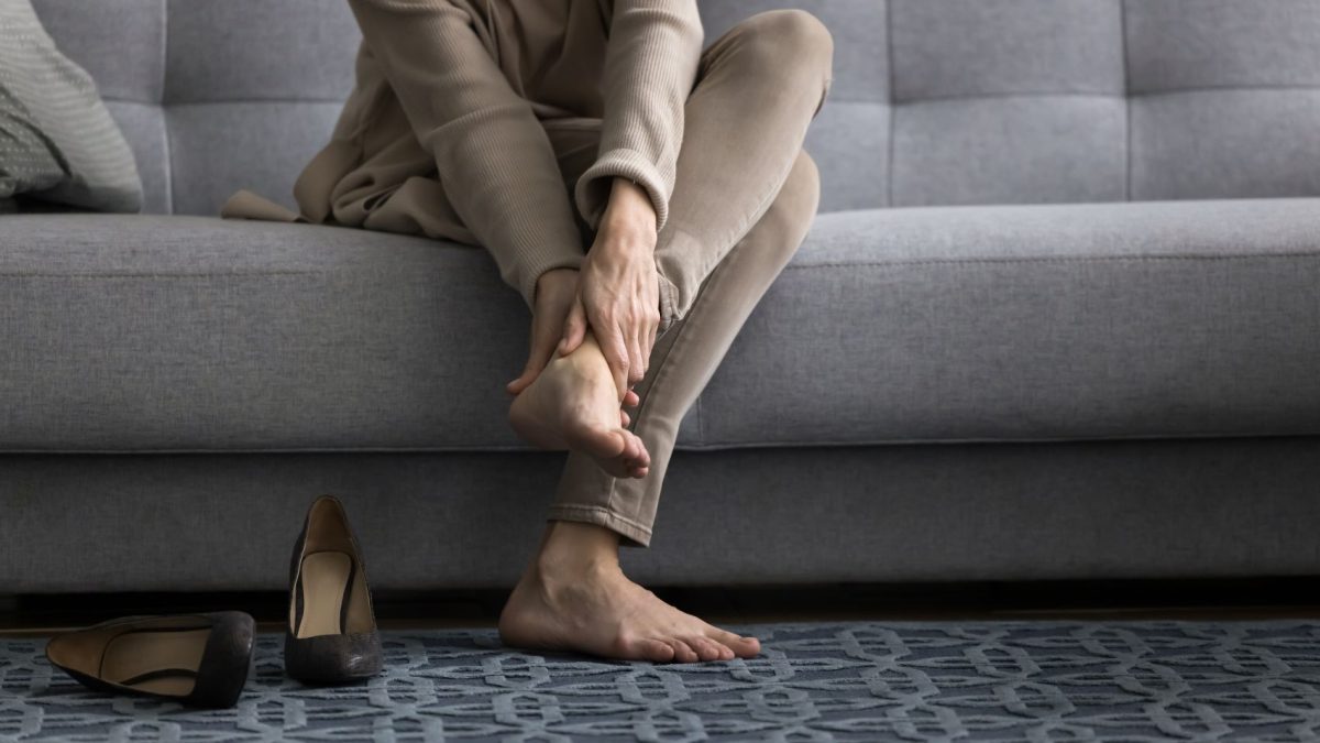 Woman sitting on a couch, rubbing her foot, with high heels placed on the floor nearby, highlighting comfort needs for long hours.