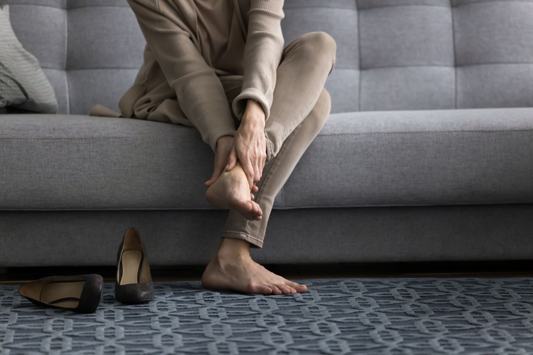 Woman sitting on a couch, rubbing her foot, with high heels placed on the floor nearby, highlighting comfort needs for long hours.