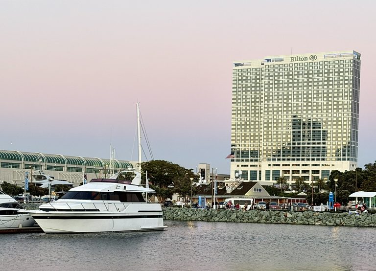 Hilton San Diego Bayfront hotel beside a marina with yachts, set against a pastel sunset sky.