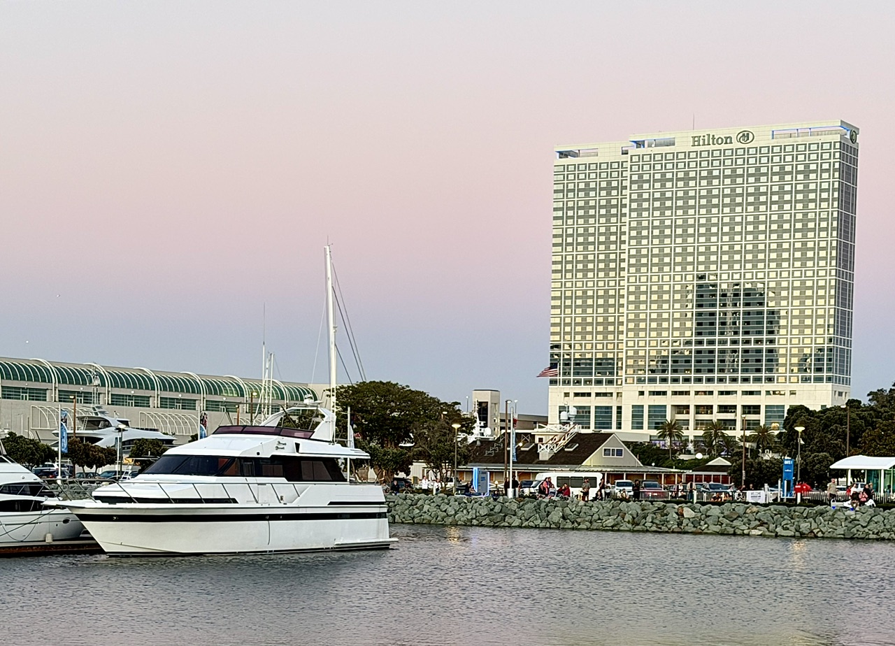 Hilton San Diego Bayfront hotel beside a marina with yachts, set against a pastel sunset sky.