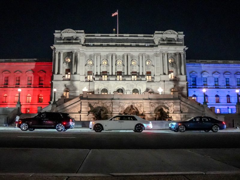 Three Rolls-Royce cars parked in front of a grand building illuminated in red, white, and blue for America's 250th anniversary.