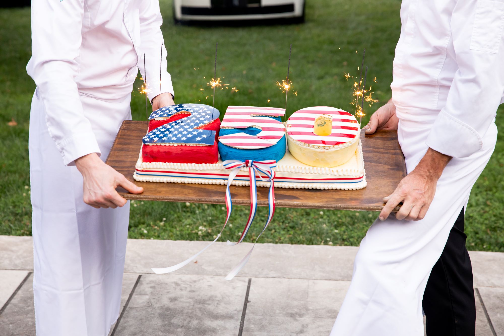 Chefs hold a large celebratory cake shaped like "250," decorated in patriotic colors and sparklers.