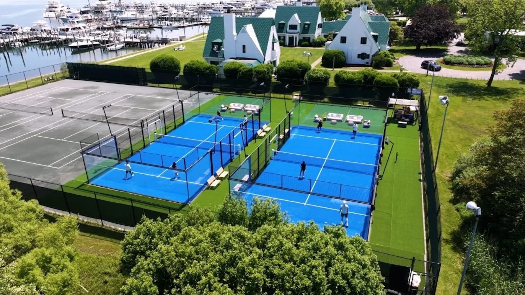 Aerial view of luxury padel courts surrounded by greenery and waterfront, with players actively engaging in the sport.