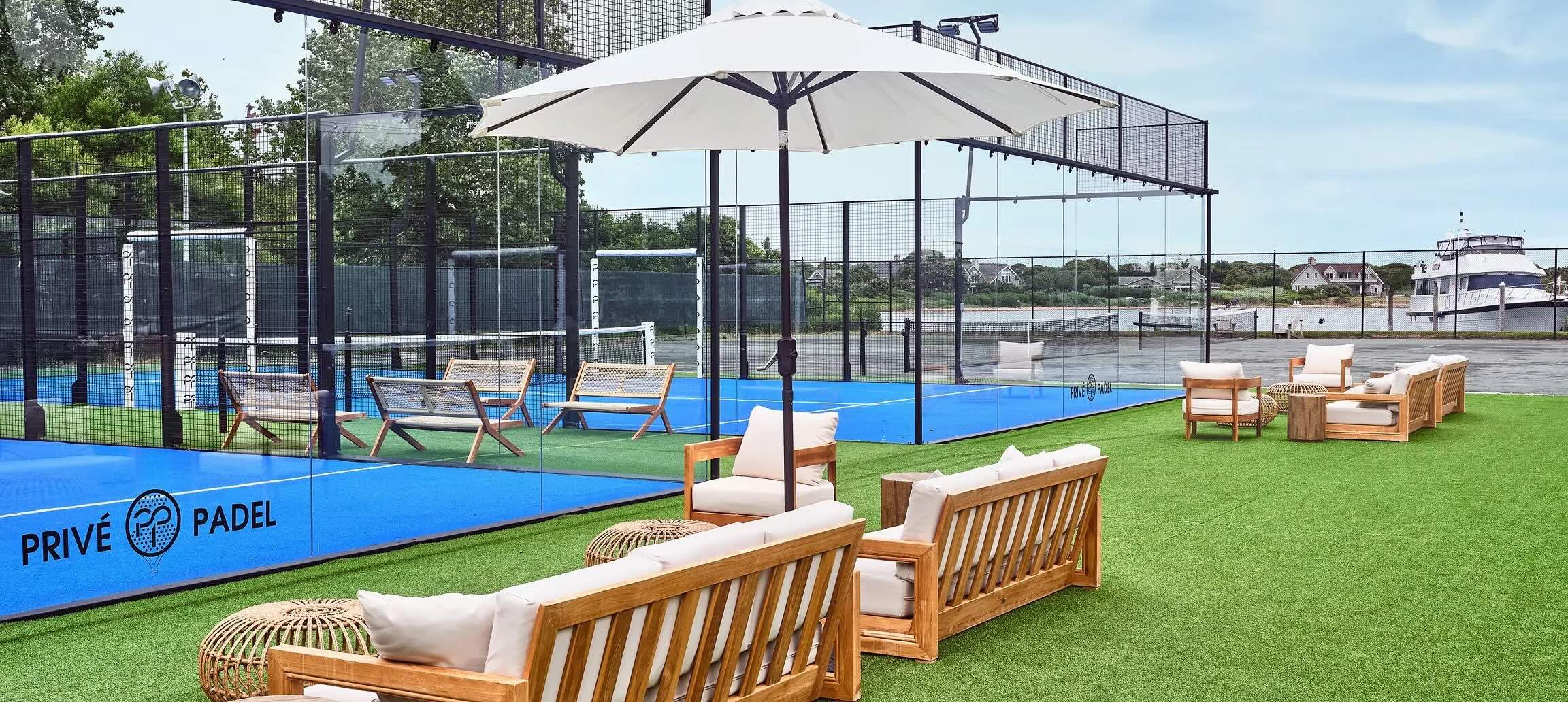 Luxury padel tennis court by Privé Padel, featuring stylish seating, an umbrella, and a scenic backdrop of water and greenery.