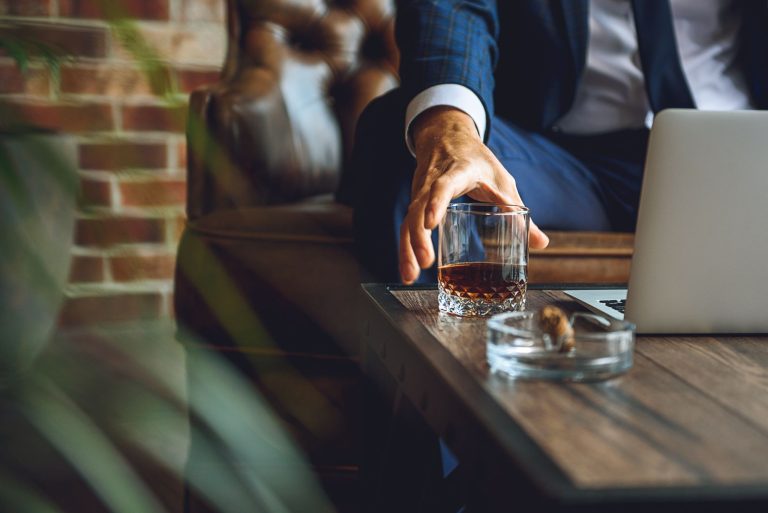 A man reaches for a glass of whiskey next to a laptop on a wooden table in a luxurious cigar lounge.