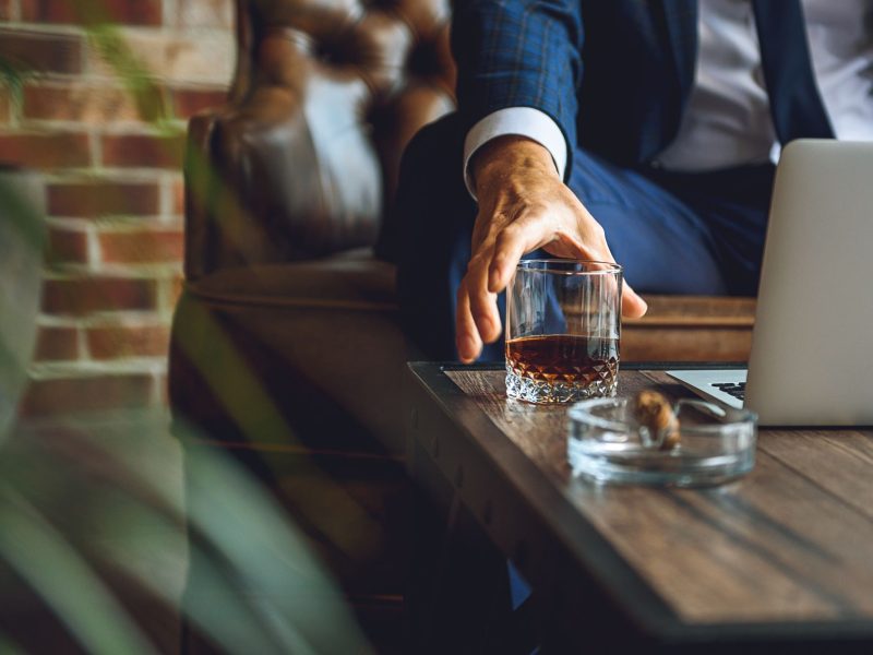 A man reaches for a glass of whiskey next to a laptop on a wooden table in a luxurious cigar lounge.
