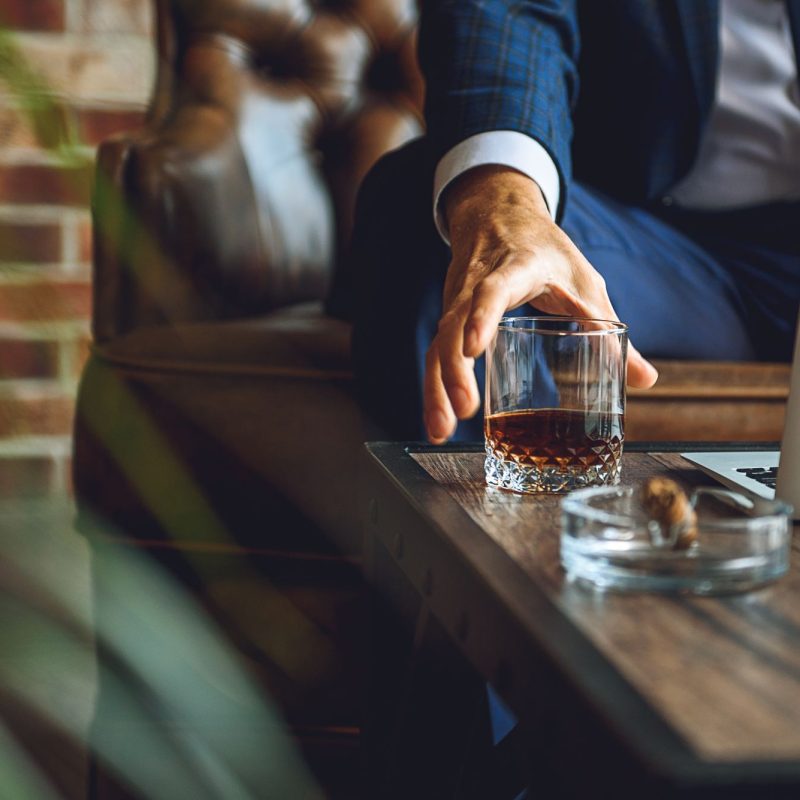 A man reaches for a glass of whiskey next to a laptop on a wooden table in a luxurious cigar lounge.