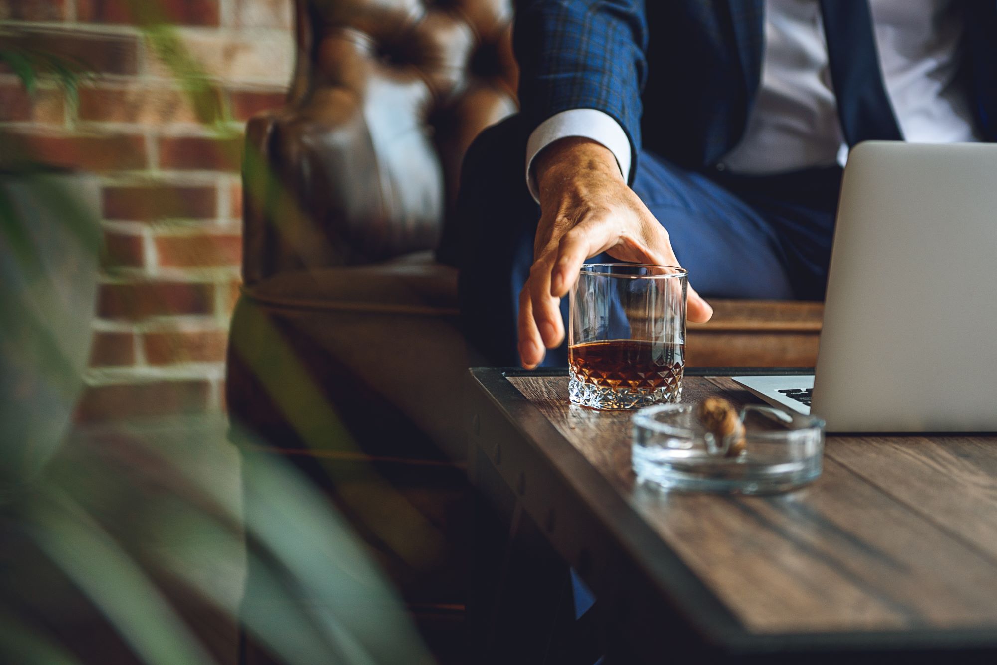 A man reaches for a glass of whiskey next to a laptop on a wooden table in a luxurious cigar lounge.