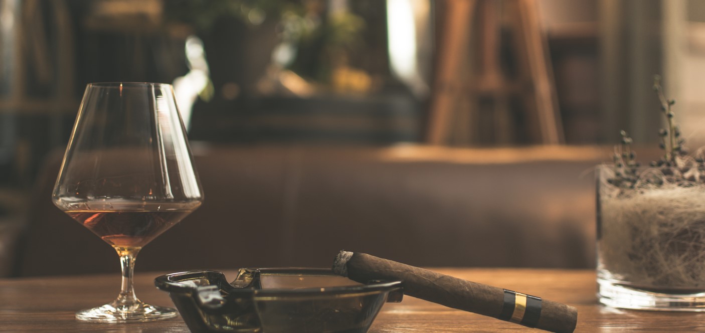 A glass of whiskey next to an ashtray with a cigar and a decorative plant in a luxurious lounge setting.