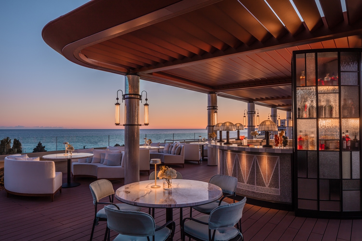 Stylish bar area with tables overlooking the ocean at sunset, featuring elegant lighting and modern decor.