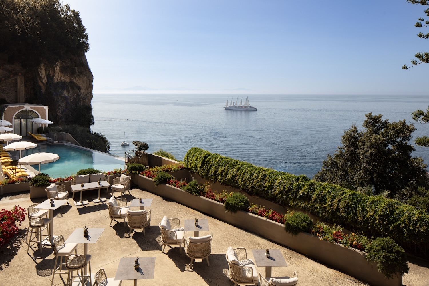 Terrace pool bar with seating and views of the ocean and a sailboat, surrounded by greenery and vibrant flowers.