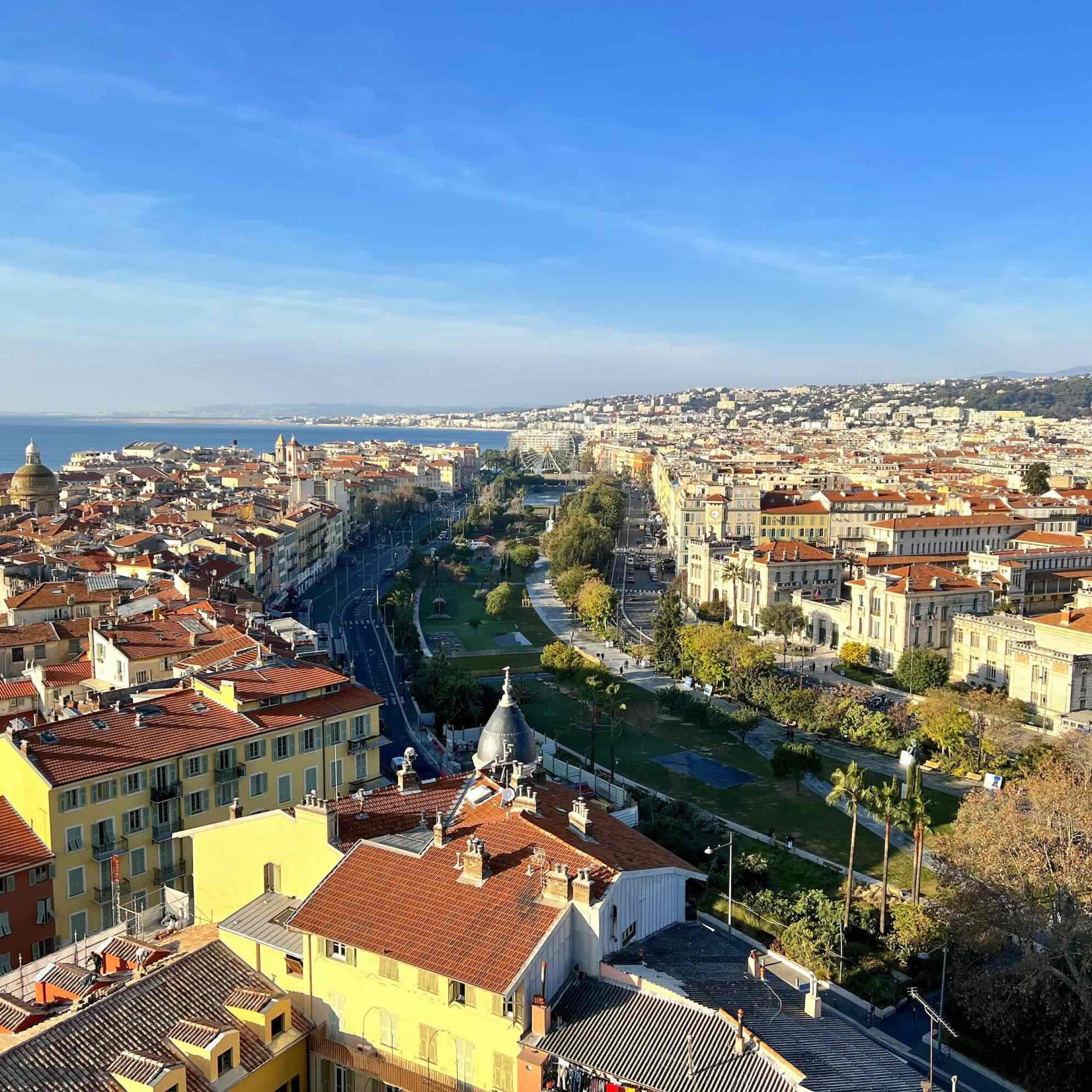 Panoramic view of Nice, France, showcasing the coastline, rooftops, and lush greenery along the promenade.