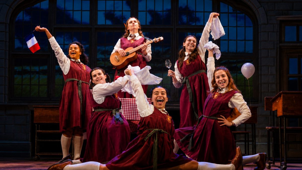 Six girls in red school dresses celebrate on stage, joyfully holding props like a guitar, a flag, and napkins during a performance.