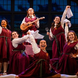 Six girls in red school dresses celebrate on stage, joyfully holding props like a guitar, a flag, and napkins during a performance.