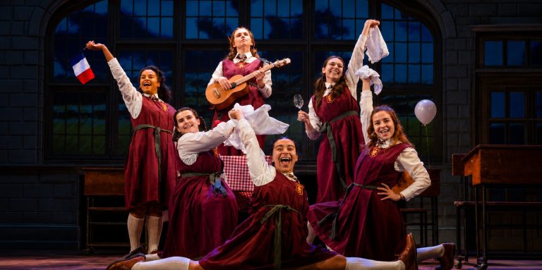 Six girls in red school dresses celebrate on stage, joyfully holding props like a guitar, a flag, and napkins during a performance.