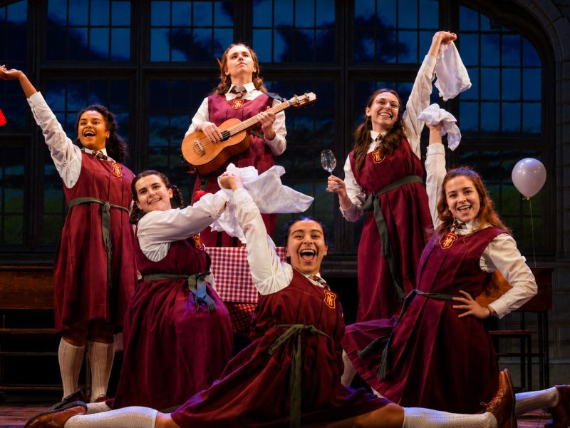 Six girls in red school dresses celebrate on stage, joyfully holding props like a guitar, a flag, and napkins during a performance.