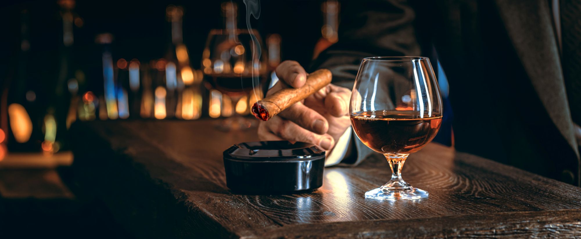 A hand holds a lit cigar above an ashtray, with a glass of whiskey resting on a wooden table in a luxurious lounge setting.