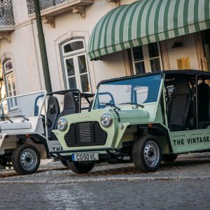 Two electric MOKE vehicles parked on a cobblestone street near a building with a green awning.