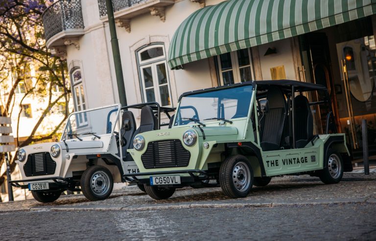 Two electric MOKE vehicles parked on a cobblestone street near a building with a green awning.