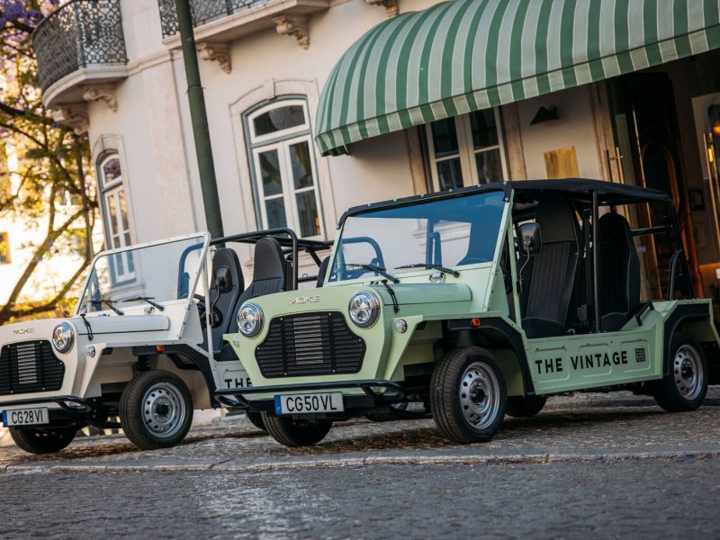 Two electric MOKE vehicles parked on a cobblestone street near a building with a green awning.