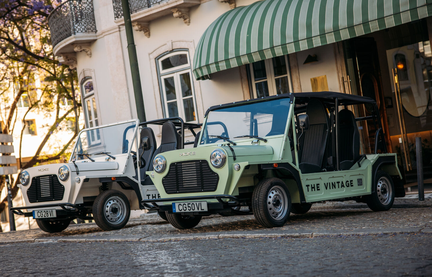 Two electric MOKE vehicles parked on a cobblestone street near a building with a green awning.