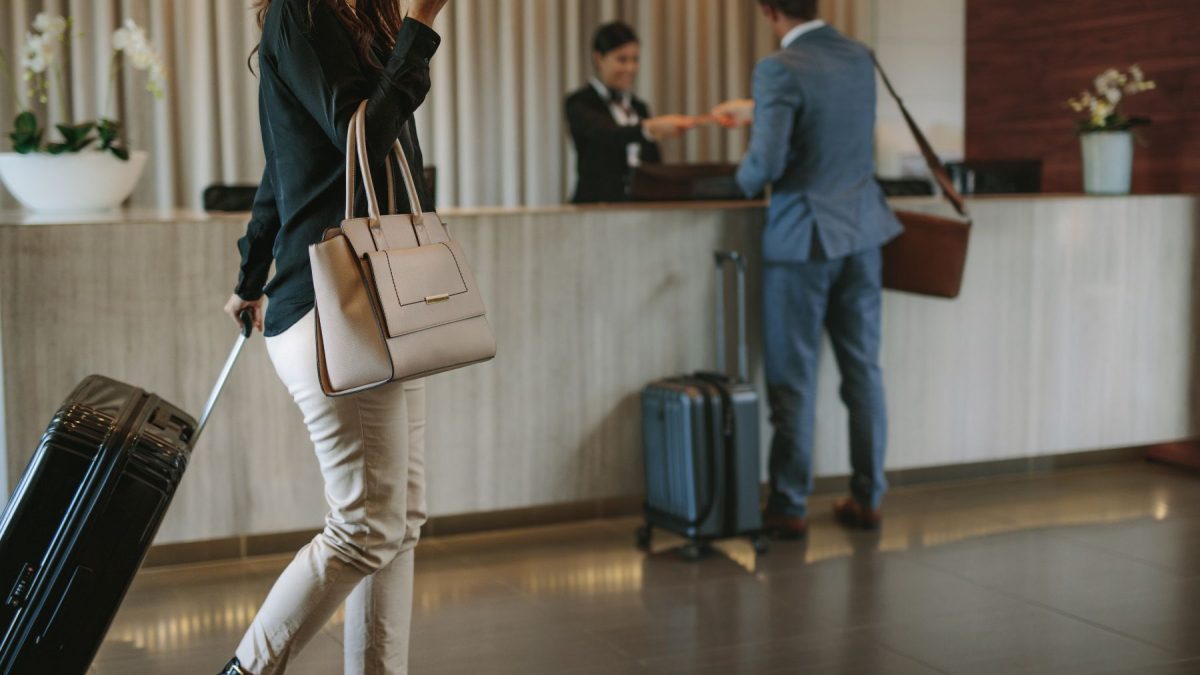 Woman checking in at a hotel with a suitcase and handbag, while a staff member assists another guest at the front desk.