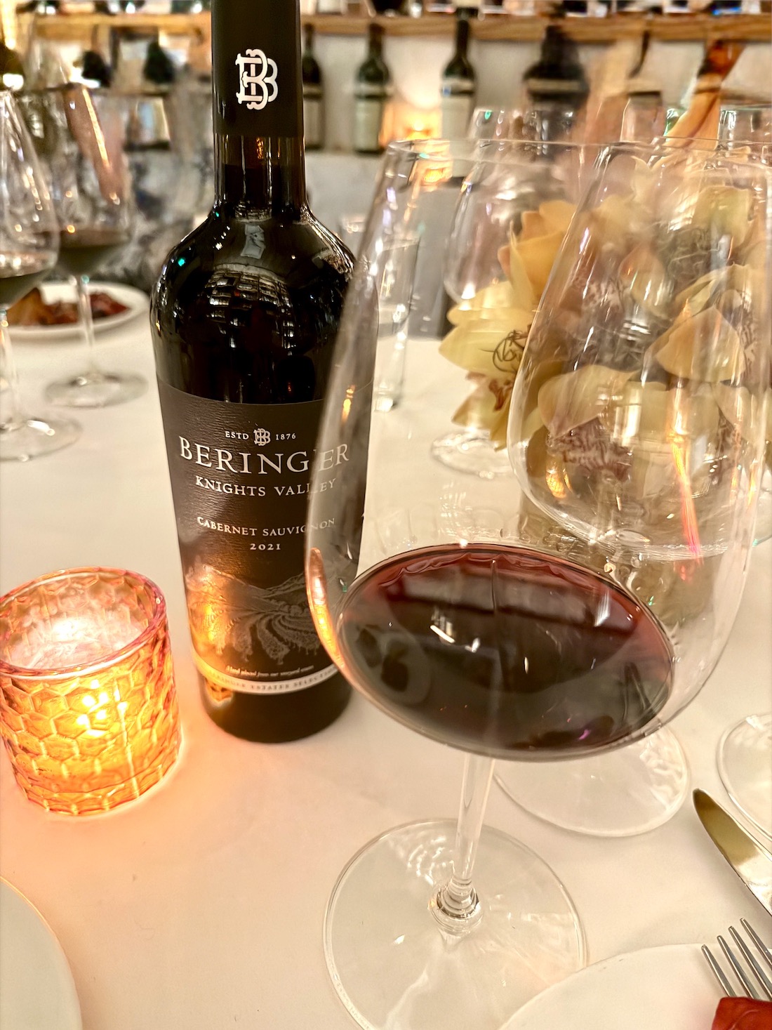 Beringer Knights Valley Cabernet Sauvignon 2021 bottle next to a filled wine glass and a glowing candle on a dining table.