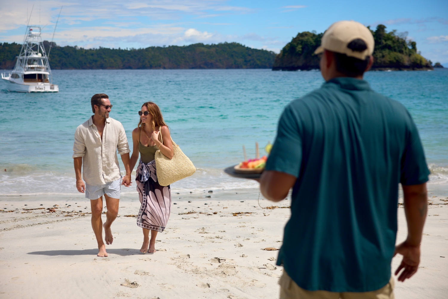 Couple walking on a sandy beach, smiling, while a server brings refreshments on a platter in the background.