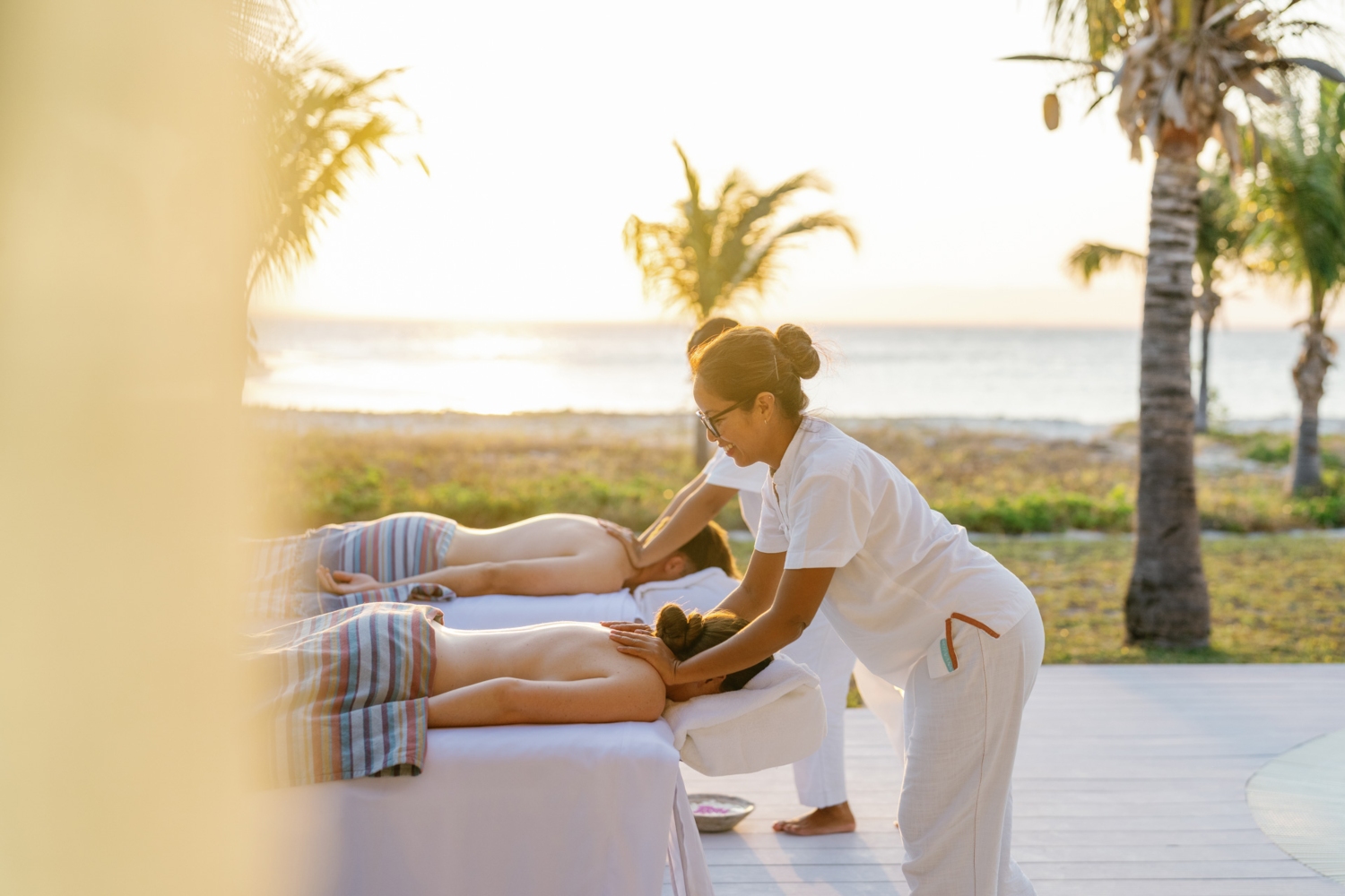 A therapist giving a massage to a client on a beachside terrace during sunset, with palm trees in the background.