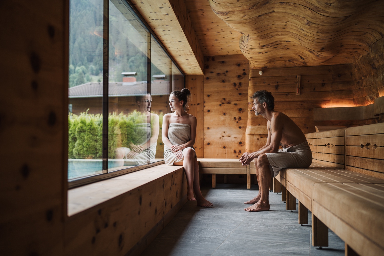 A couple relaxes in a wooden sauna, sitting on a bench by a large window overlooking a serene outdoor landscape.