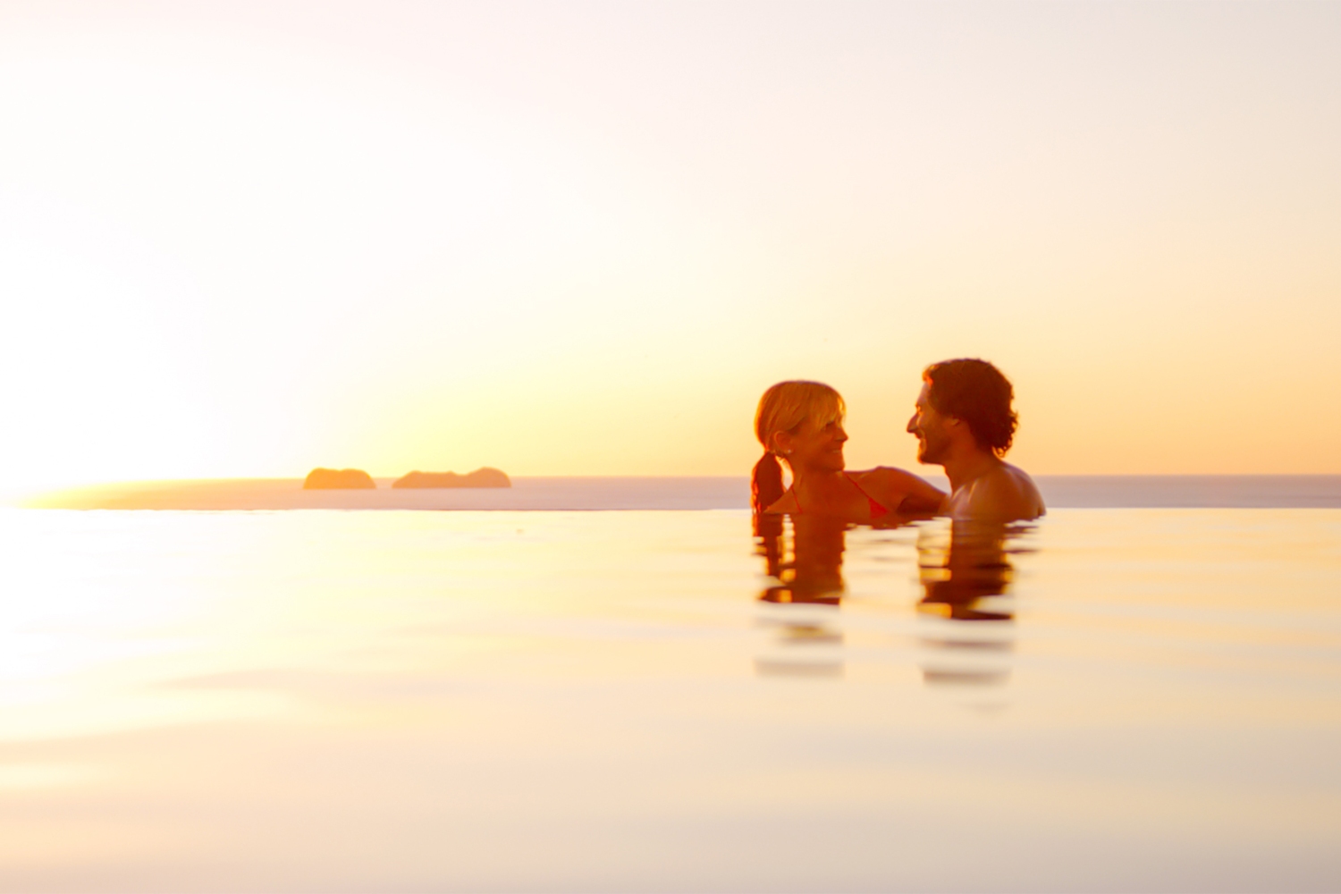 Couple enjoying a romantic moment in an infinity pool at sunset with ocean views in the background.