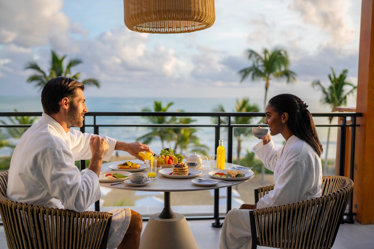 A couple enjoying a luxurious breakfast on a balcony with ocean views, surrounded by palm trees and beautiful clouds.